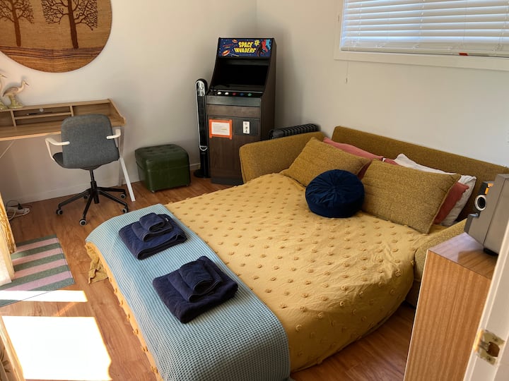 Bedroom 3 features a luxury queen bed with plush topper, luxe linen, Sheridan towels, a dedicated work desk, fan, heater, and a console packed with retro arcade classics. Sliding doors open to a sunlit deck framed by the lush garden.