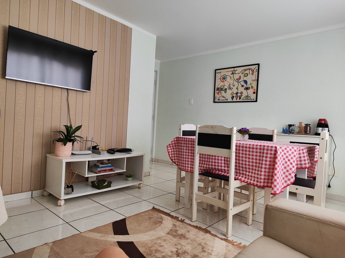 A dining area is presented with a checkered tablecloth covering a rectangular table surrounded by white chairs. A television is mounted on the wall opposite the seating area. A plant is situated on a side table, adding a touch of greenery to the space.