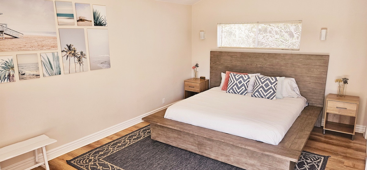 A spacious bedroom features a king-size bed with a wooden frame and a simple white duvet. Two bedside tables with lamps stand on each side. A series of coastal-themed wall art is displayed above the bed, and natural light enters through a large window.