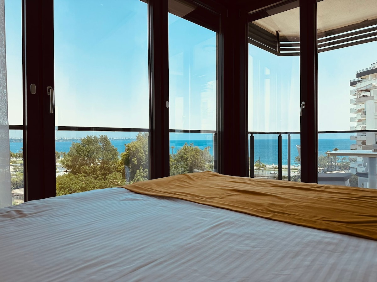 A spacious bedroom features large glass windows offering an expansive view of the sea and surrounding landscape. The room is furnished with a neatly made bed, and sunlight fills the space, creating a bright and airy atmosphere.