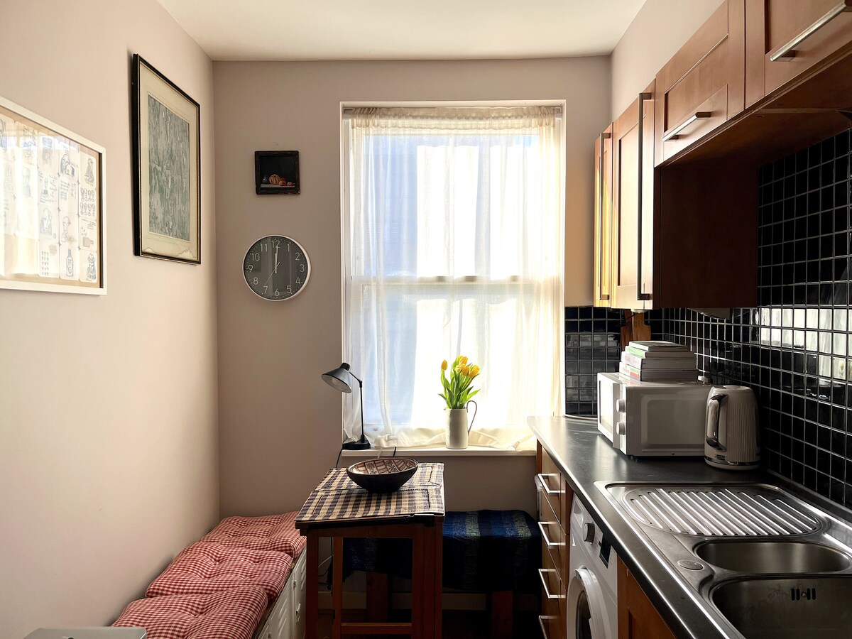A well-lit kitchen features wooden cabinetry and a countertop with a sink and a cooking area. A small round table is adorned with a decorative bowl and a plant. Natural light filters through a window, highlighting a cozy seating nook with cushions.