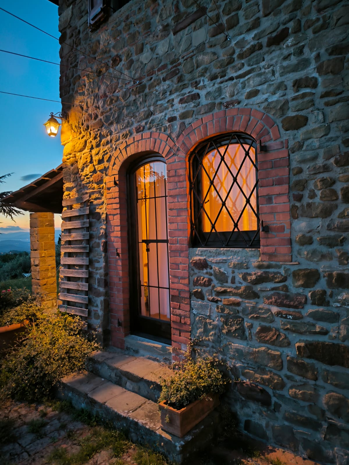 The entrance of the villa features arched brick doorways with diamond-patterned windows. Warm, orange light emanates from within, contrasting against the rustic stone facade. Surrounding greenery is visible, creating a welcoming ambiance as twilight approaches.