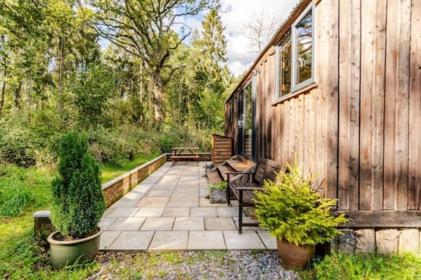 A spacious patio area features grey stone tiles, providing a natural transition between the cabin and the surrounding woodland. Wooden benches are positioned for relaxed seating, accompanied by potted plants adding greenery to the space. Tall trees frame the scene, enhancing the secluded atmosphere.