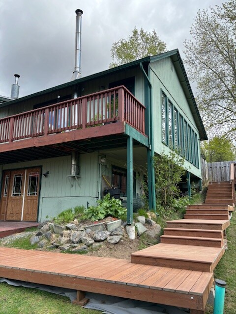 A two-story green house features large windows along its façade, allowing ample natural light. A wooden deck is visible above raised garden beds, while a set of wooden steps leads up to the entrance. The surrounding greenery adds to the outdoor appeal.