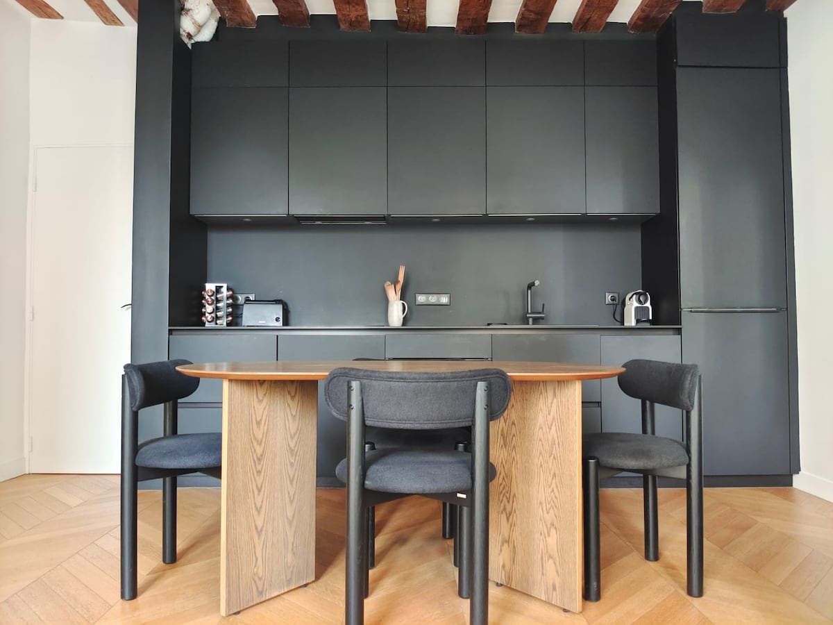 A modern kitchen area features sleek black cabinetry and a wooden dining table surrounded by four black chairs. The space is complemented by wooden beams in the ceiling and includes essential appliances, creating a functional area for dining and cooking.
