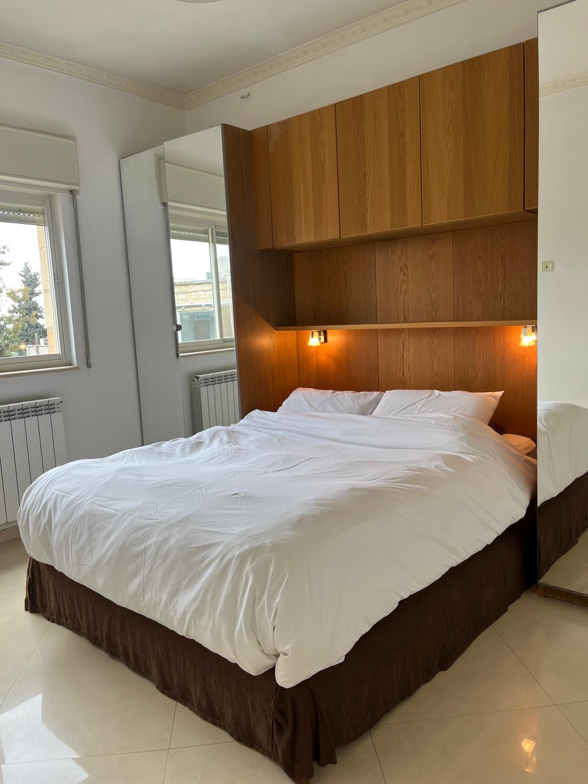 A cozy bedroom is highlighted by a spacious bed with white bedding and a dark brown bed skirt. Light wood cabinetry and two bedside lights are positioned above the bed, while large windows provide natural light, complemented by light-colored walls and ceramic flooring.