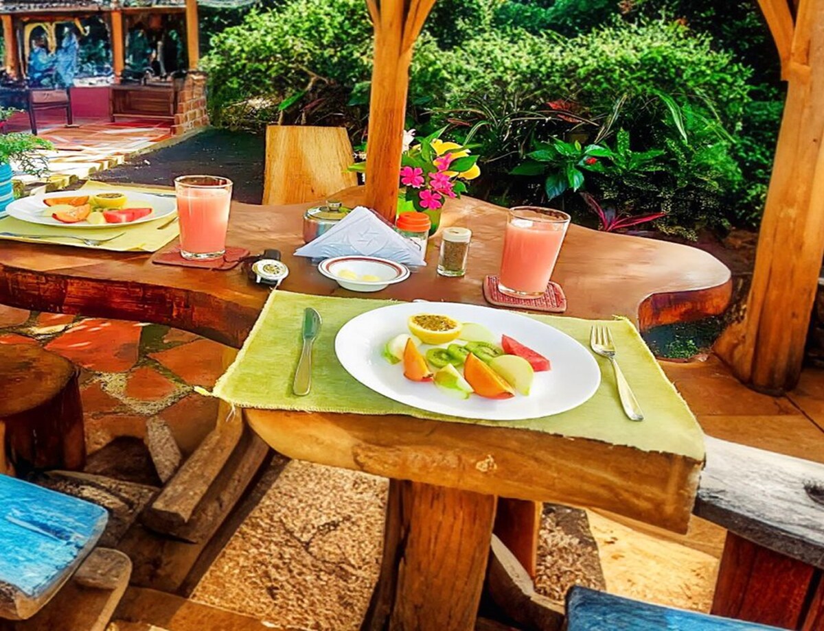 A rustic dining table is set outdoors, featuring a colorful plate of fresh fruit and two glasses of pink beverage nearby. Lush greenery and vibrant flowers surround the area, creating a serene outdoor dining space.