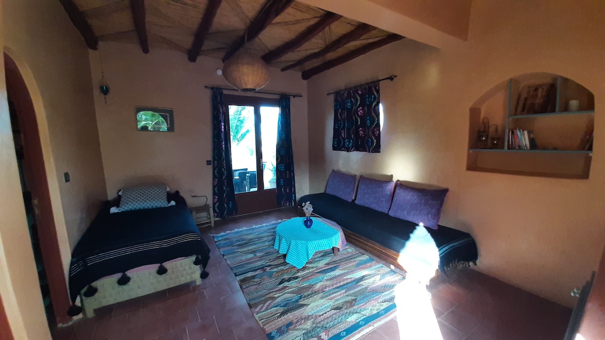 A spacious living area is equipped with two single beds and decorative cushions. A low table is centered on a patterned rug, while large windows overlook the outdoor space. Colorful curtains frame the windows, and wooden beams add character to the ceiling.