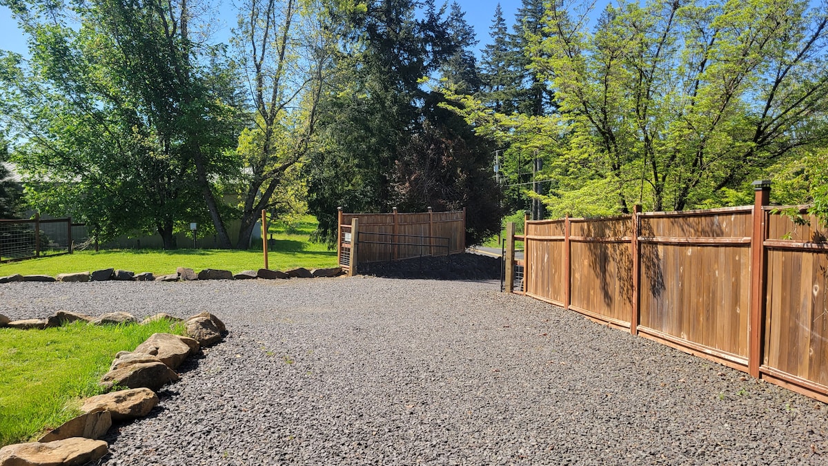 A spacious gravel driveway leads to a wooden gate, flanked by a tall wooden fence. Lush greenery and trees surround the area, creating a peaceful natural setting. The gravel surface offers ample parking space, providing easy access to the cottage.