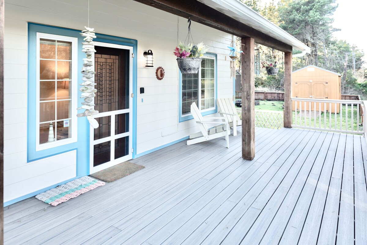 The front porch features wooden decking and inviting seating, including two white chairs. A decorative mobile hangs nearby, and potted plants are positioned at the entrance. The door is framed in blue, adding a touch of color to the entryway.