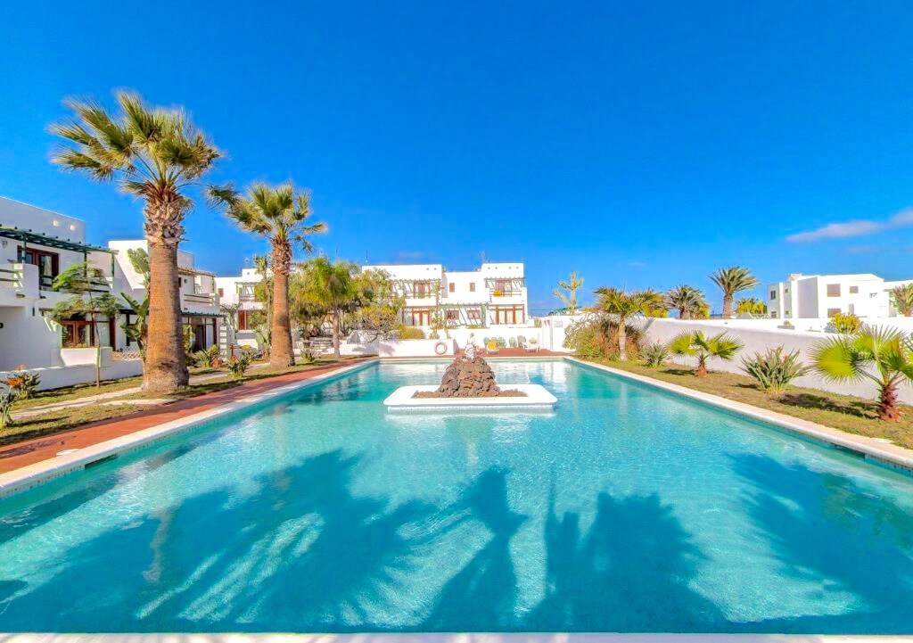 A serene pool area is highlighted, surrounded by palm trees and lush greenery. The clear blue water reflects the sky, while the landscape features well-maintained gardens and decorative elements. White buildings can be seen in the background, contributing to the relaxed atmosphere.
