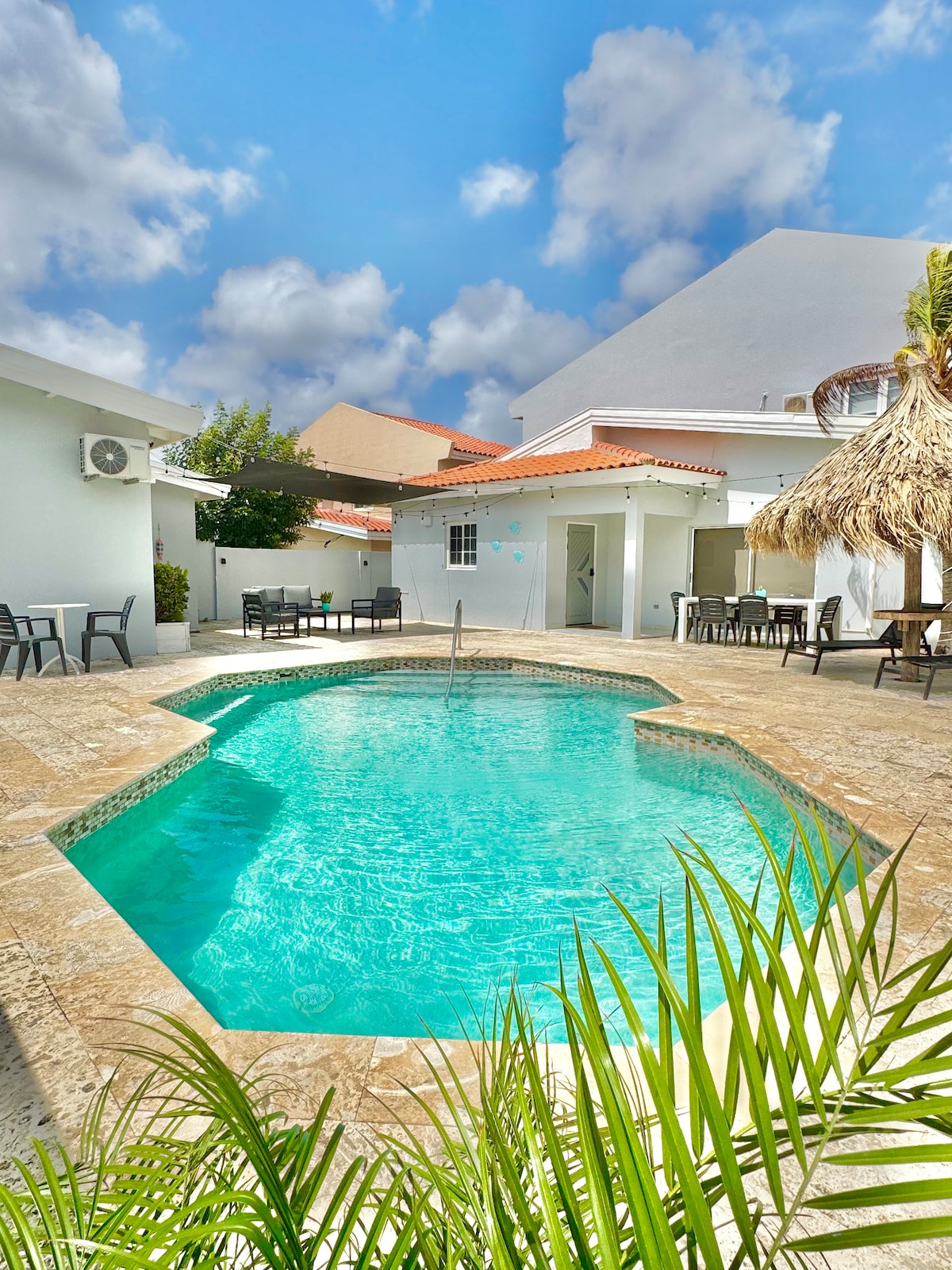 A serene private pool area is seen, featuring clear turquoise water surrounded by a natural stone terrace. Shaded seating areas, including a thatched-roof gazebo, are positioned nearby, set against a backdrop of soft clouds and bright blue skies.