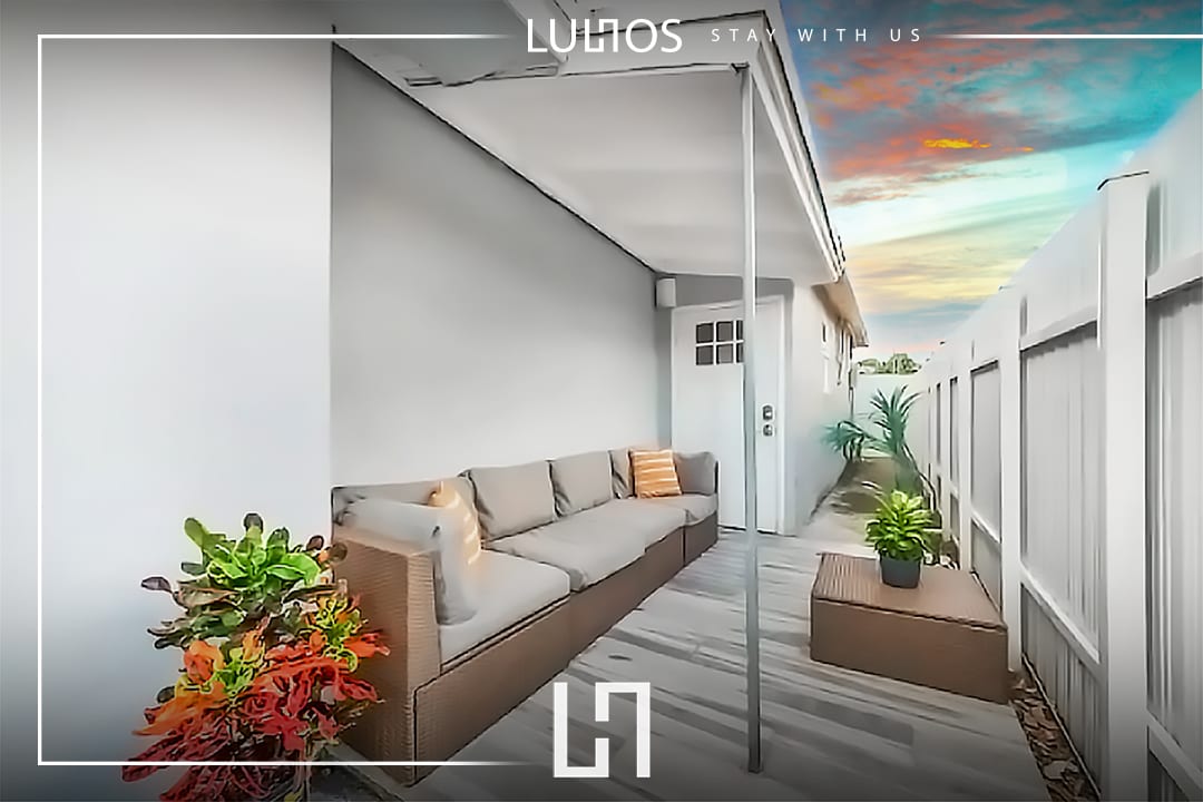 An outdoor seating area is displayed with a comfortable couch positioned against a wall. Potted plants with vibrant foliage add color, while a pathway leads to a door. A peaceful sky with soft hues can be observed in the background.