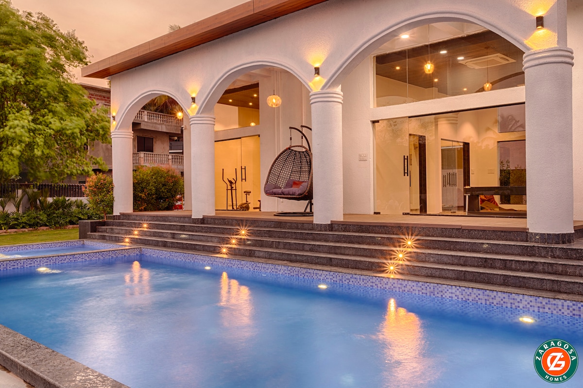 A swimming pool is set against a backdrop of lush greenery and a spacious property. The pool features clear blue water, reflecting the surrounding landscape. Steps lead down to the water's edge, providing easy access. The scene is enhanced by warm sunset lighting.