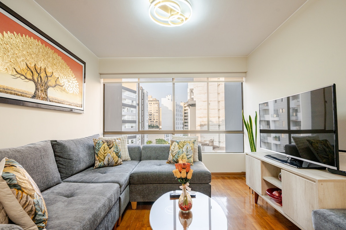 A spacious living area features a gray sectional sofa adorned with decorative cushions. A round glass coffee table holds a vase of fresh flowers. Large windows frame views of the city, while a flat-screen TV is positioned on a light wooden console. Modern lighting adds a soft glow.