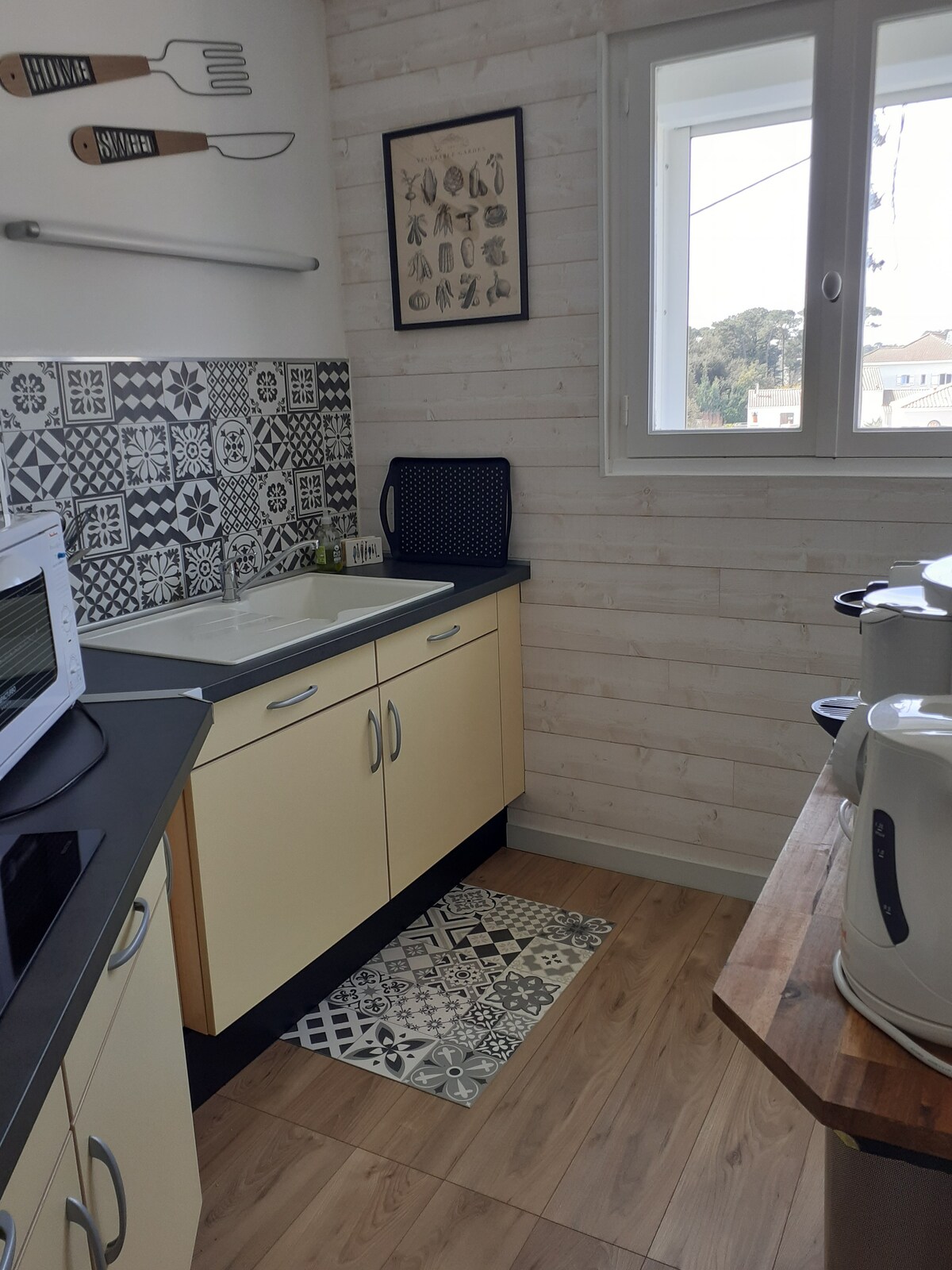 A well-equipped kitchen features a sink, microwave, and countertop appliances. Decorative tiles adorn the wall, contrasting with the light cabinetry. A small rug lies on the floor, and a window allows natural light to brighten the space.