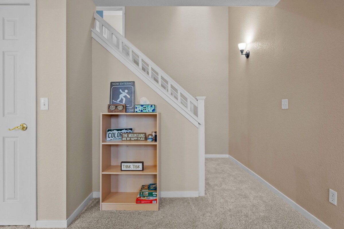 An interior space features soft beige walls and plush carpeting. A small bookshelf with various decor items and games stands against the wall, while a staircase with white railings leads to the upper floor. A light fixture illuminates the area.