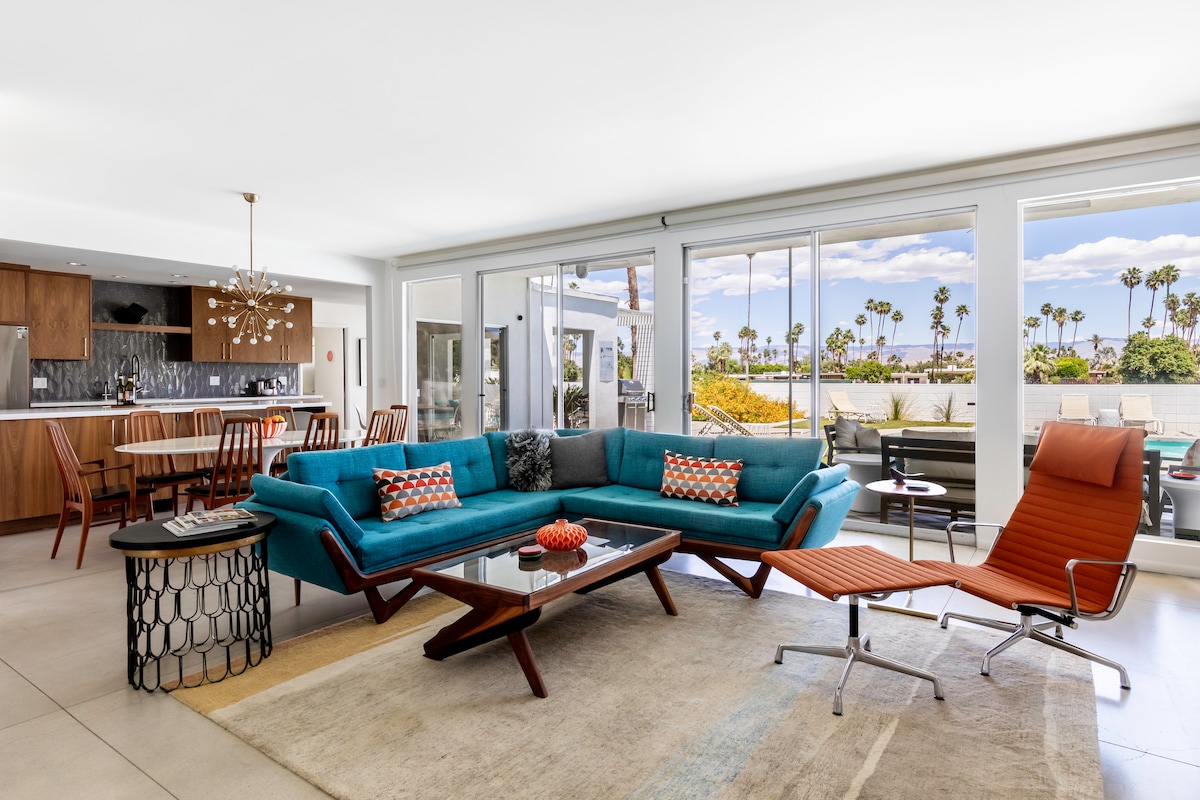 An open-concept living area features a large teal sofa with decorative pillows, a stylish coffee table, and an orange lounge chair. Floor-to-ceiling glass doors allow natural light to fill the space, offering a view of palm trees and outdoor dining. The modern kitchen area is visible in the background.
