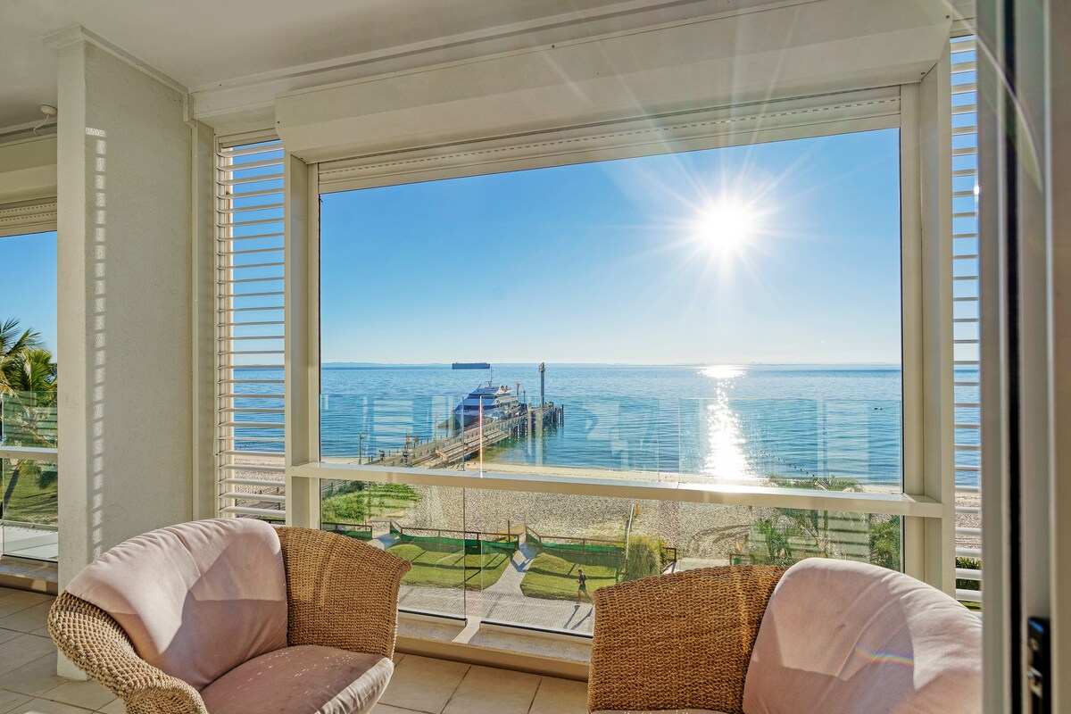 The image features a sunlit room with large windows overlooking the beach and Moreton Bay. Two comfortable chairs are positioned against the glass, allowing for unobstructed views of the water and pier. The sunlight reflects on the water, creating a bright and inviting atmosphere.