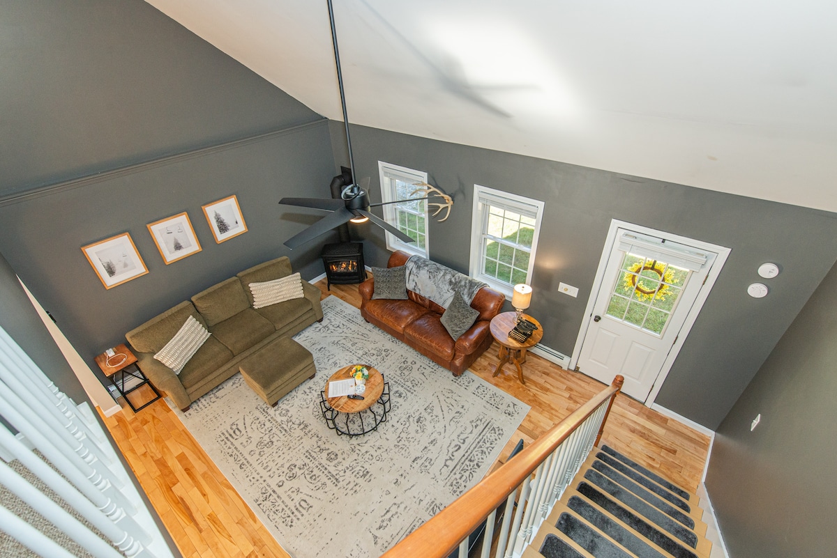 An open living area is visible from above, featuring a cozy sectional sofa and a leather armchair arranged around a circular coffee table on a patterned area rug. Large windows allow ample natural light while a ceiling fan adds to the inviting ambiance.