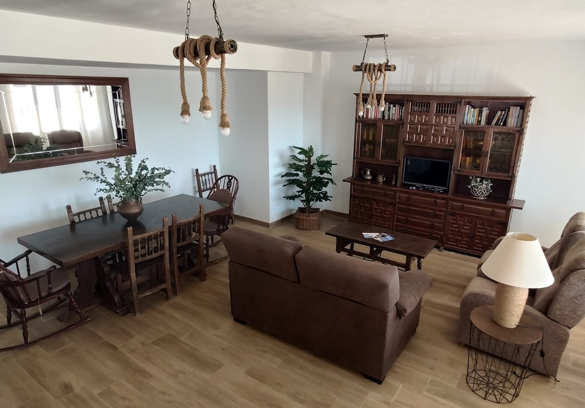 An open living space features a combination of wooden furniture, including a dining table and chairs, alongside a comfortable brown sofa. A decorative shelving unit holds books and a television, while plants add a touch of greenery. Natural light fills the area through large windows.