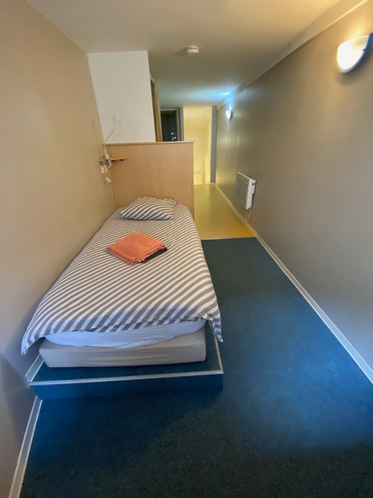 A compact sleeping area features a single bed with a striped coverlet, positioned against a light-colored wall. An orange towel is placed on the bed. The space is illuminated with soft lighting, and blue carpeting covers the floor, creating a cozy atmosphere.