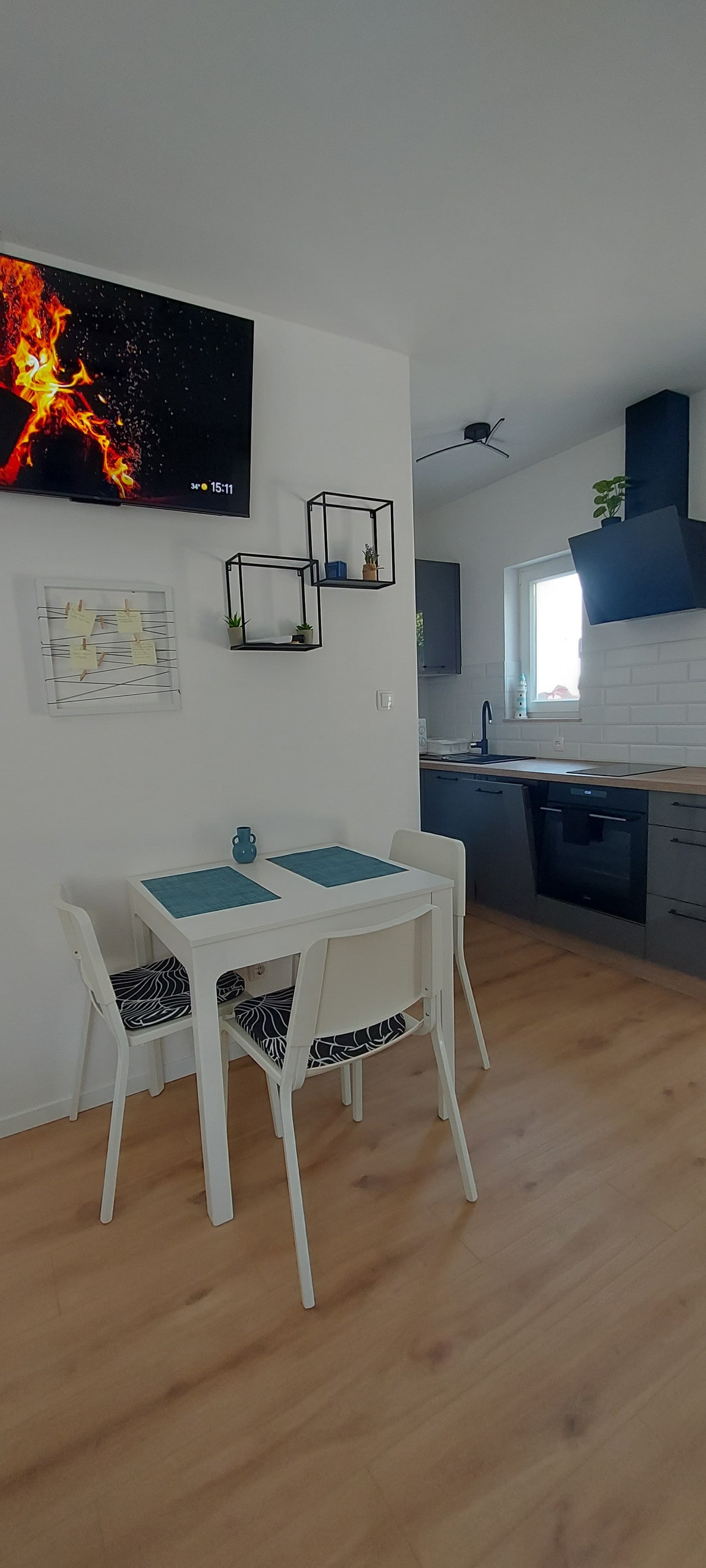 A dining area features a small table with two white chairs, positioned next to a modern kitchen. A television set is mounted on the wall, above a decorative framed piece. Natural light filters through a window, illuminating the light wood flooring and sleek appliances.