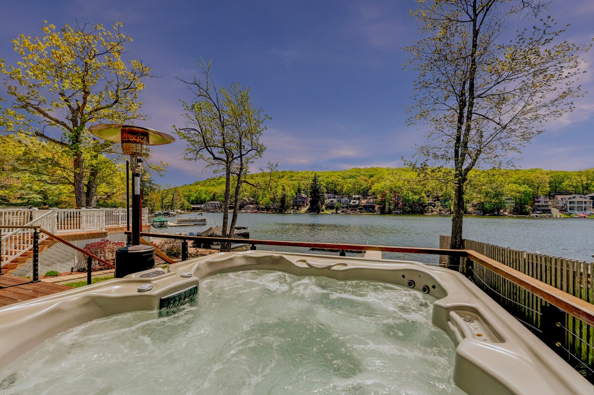 A spacious Viking hot tub sits on a wooden deck, with bubbling water visible. In the background, a serene lake reflects lush green foliage, creating a tranquil setting. Surrounding trees offer shade, while a patio heater provides warmth for cooler evenings.