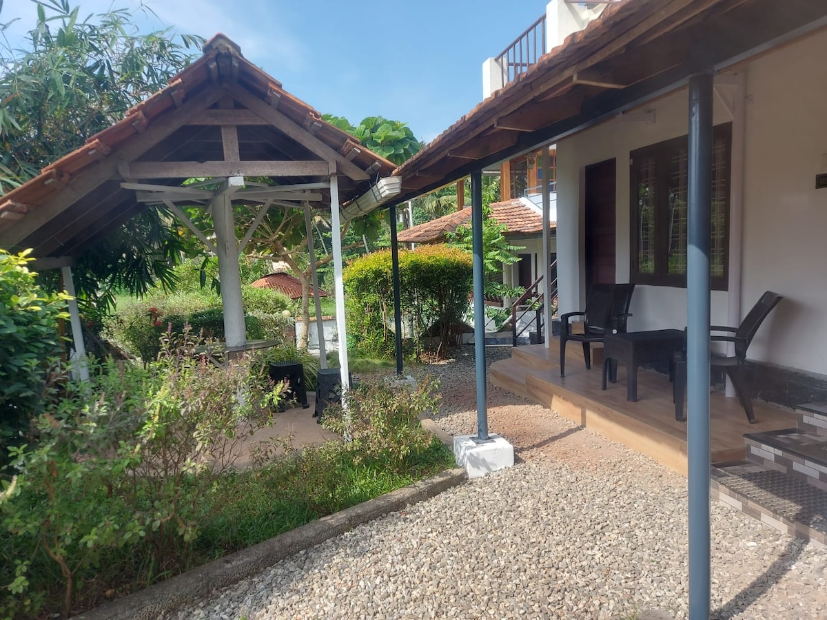 A charming outdoor area features a wooden gazebo surrounded by lush greenery. The pathway leads to a spacious porch with two chairs, providing a serene space to relax. The natural stone and gravel landscaping enhance the tranquil ambiance of this inviting setting.