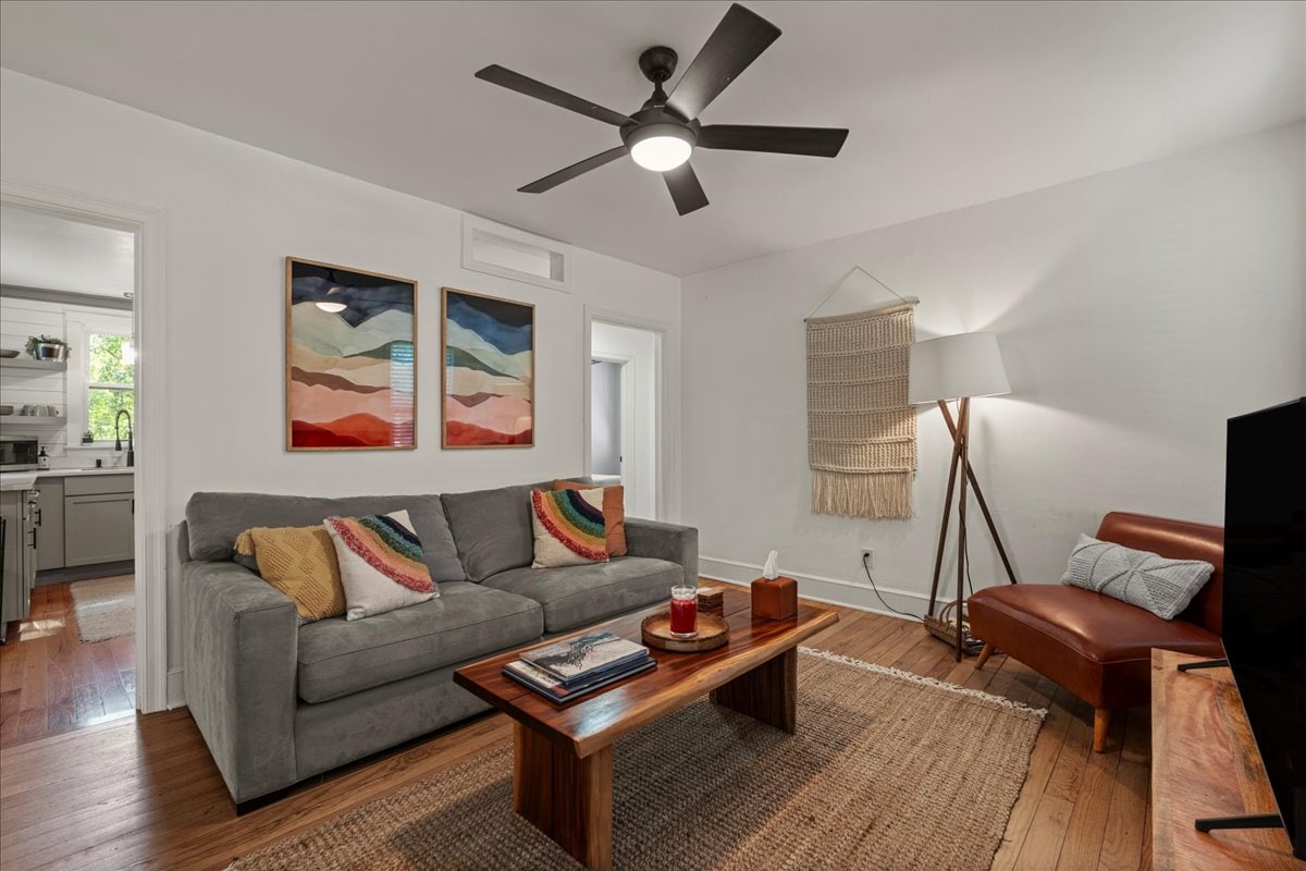 A comfortable living area showcases a gray sofa adorned with colorful pillows. A wooden coffee table is positioned in front, holding books and decorative items. Two abstract wall art pieces add visual interest, while a ceiling fan and a floor lamp provide illumination, enhancing the welcoming ambiance.