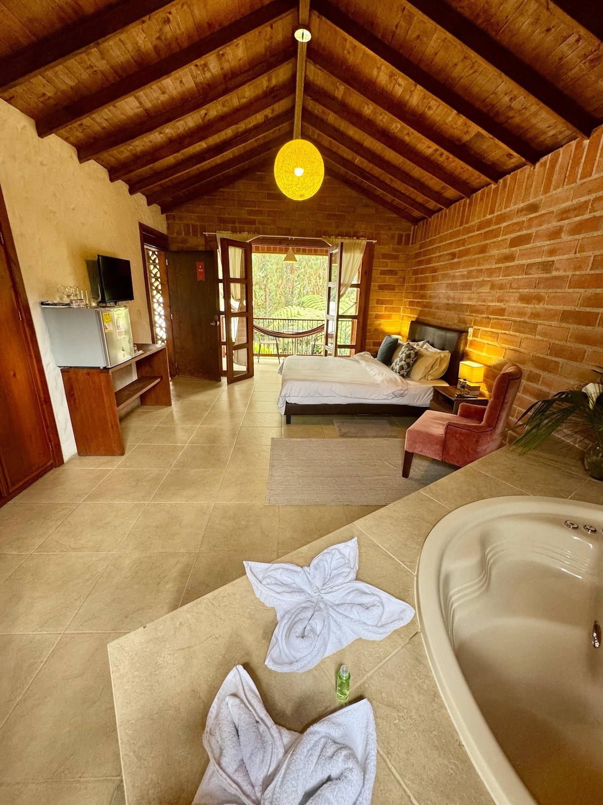 An interior view of a spacious room is displayed, featuring a double bed with a plush headboard. A cozy armchair is positioned next to the bed, while a small TV stand and mini-fridge are visible. Natural light enters through double doors, revealing a lush outdoor view.