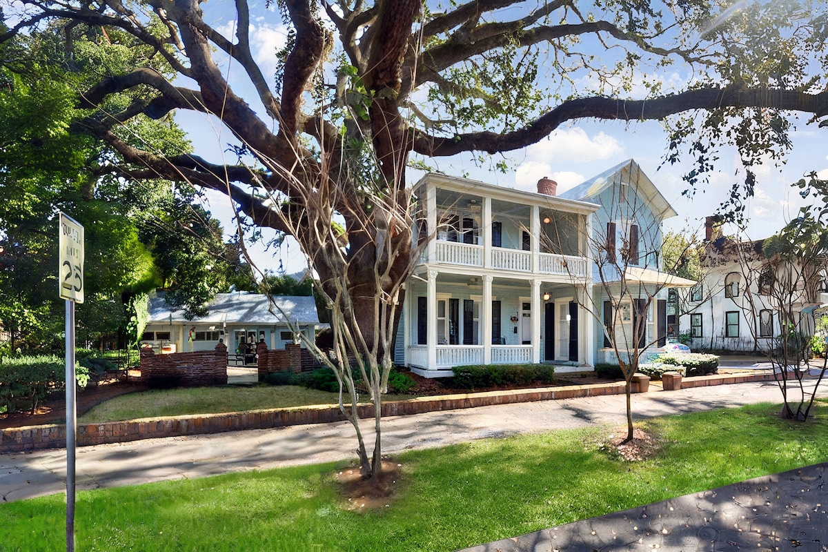 A charming two-story cottage is nestled among lush greenery and mature trees. The front porch features a welcoming railing, while large windows allow natural light to fill the interior. The inviting facade is complemented by a well-maintained lawn and surrounding garden area.