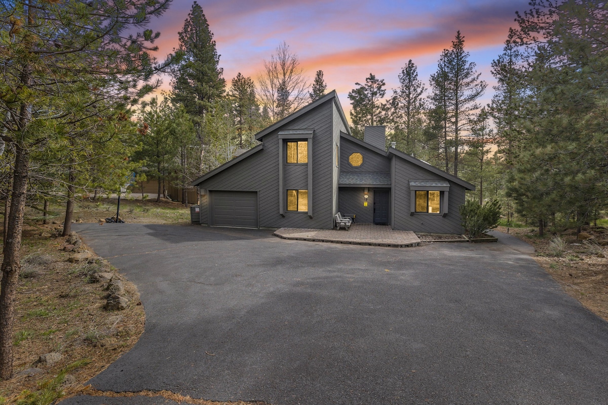 The modern home is surrounded by tall trees, featuring a contemporary design with a dark exterior. A spacious driveway leads to the entrance, and large windows reflect the evening sky, offering a glimpse of the tranquil surroundings.