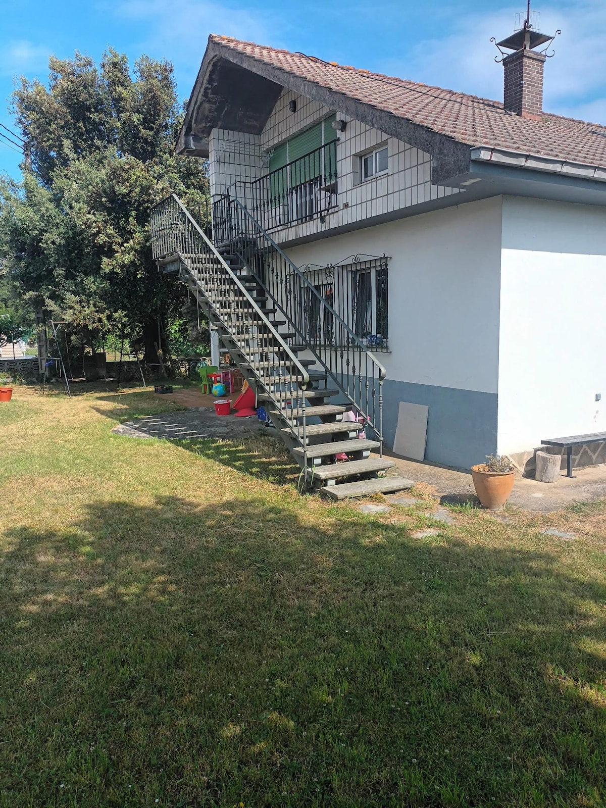 An exterior view of the house showcases a stone and iron staircase leading to the upper floor. Lush green grass surrounds the base of the property, complemented by trees in the background. Outdoor seating can be seen to the side, creating an inviting space for relaxation.