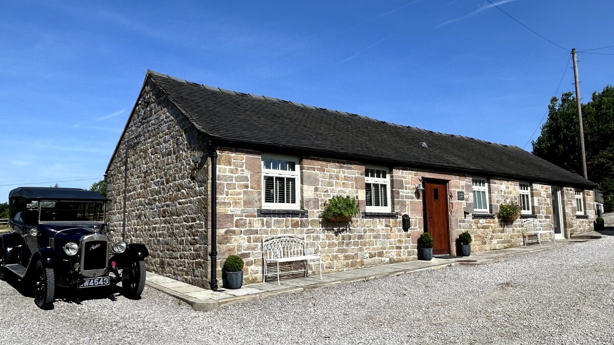 An elegant stone barn conversion is showcased under a clear blue sky. The exterior features a well-maintained façade with traditional elements, framed by neatly kept flower pots. A vintage car is parked in front, adding to the character of the setting.