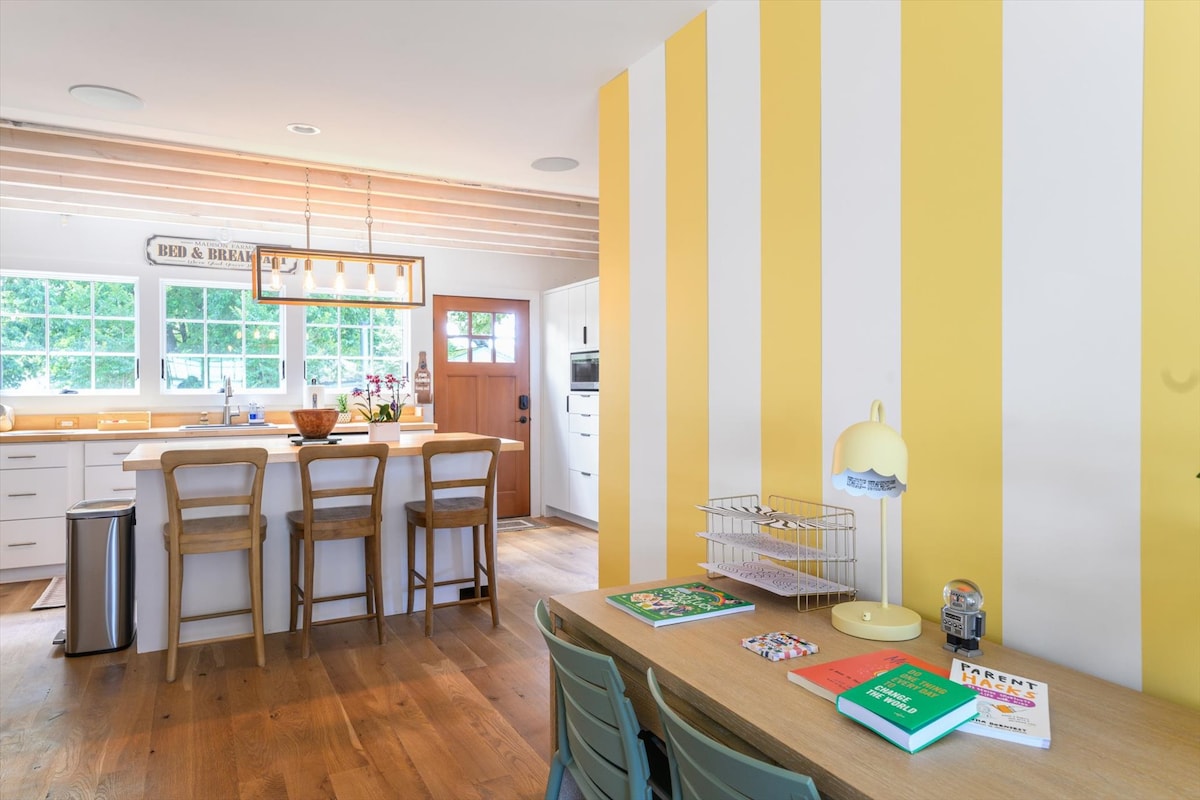 The kitchen and dining area feature cheerful yellow striped walls and wooden flooring. A large island with seating is visible, alongside a light wooden dining table with chairs. Cookware and decorative items are present, enhancing the functional layout. A friendly atmosphere is conveyed by natural light from the windows.