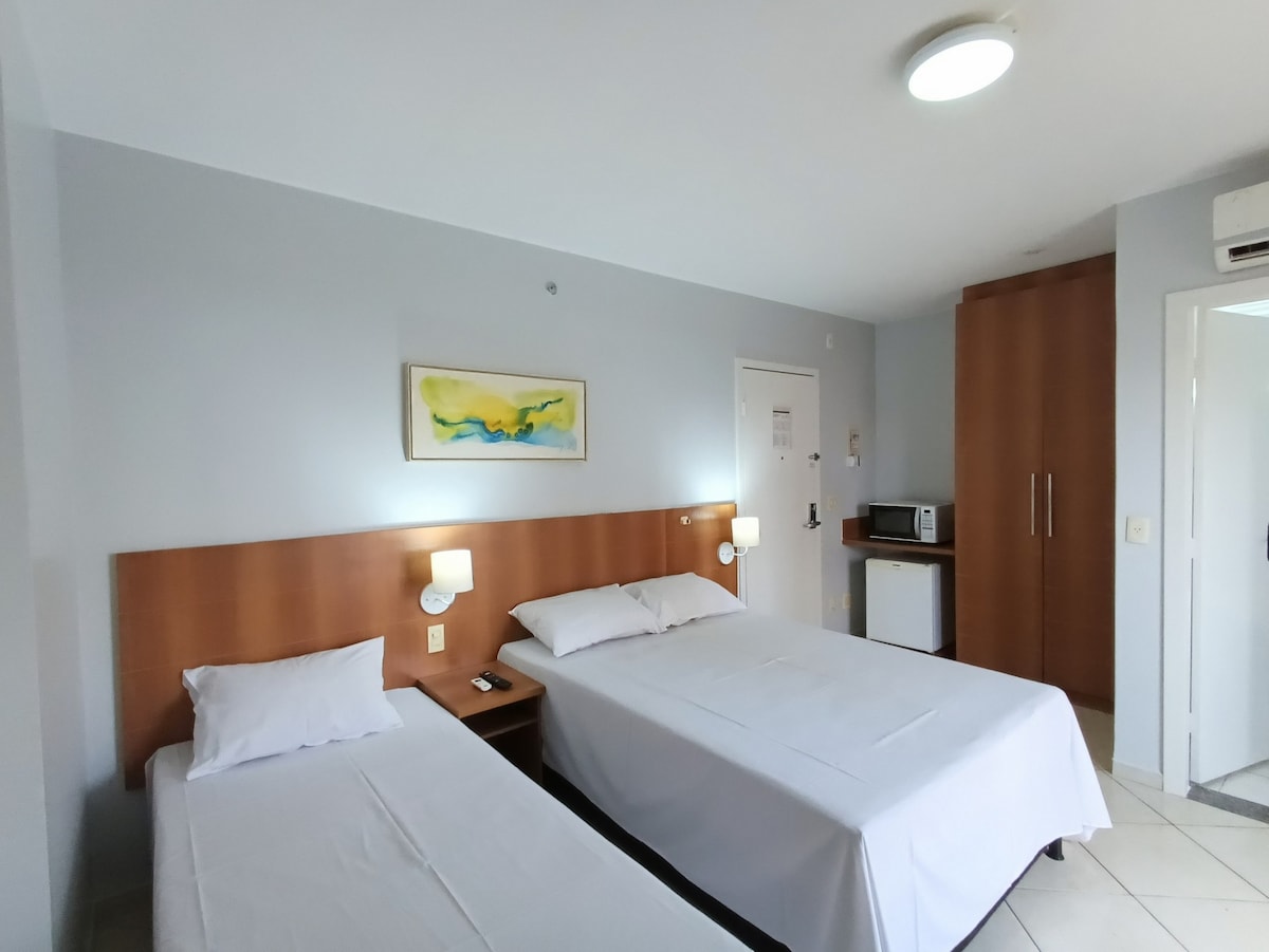 A room features a spacious double bed and a single bed, both with crisp linens. Warm light is provided by bedside lamps on either side of the beds. A kitchenette with a microwave and refrigerator is visible, along with a closet and an entry door.