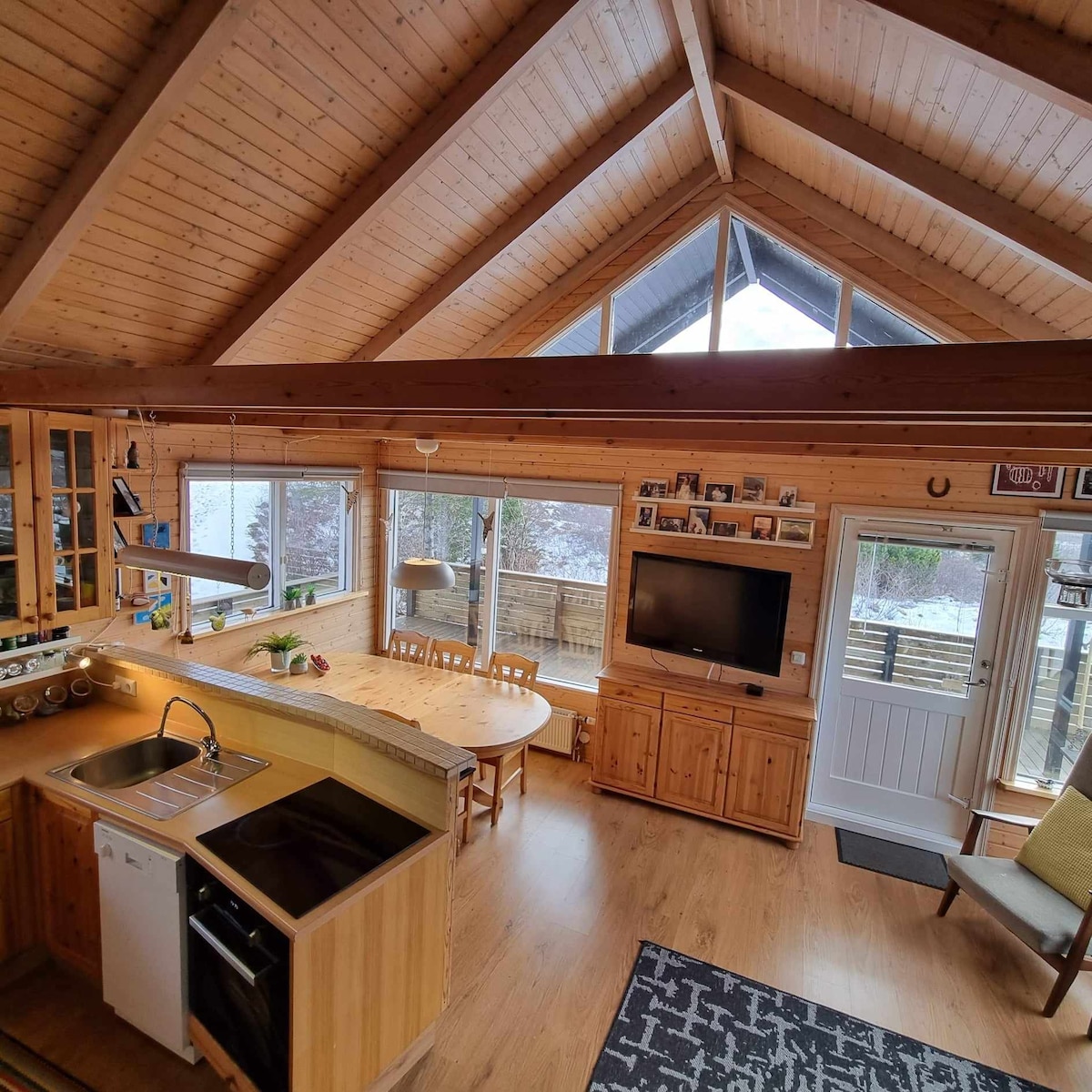 An open kitchen area features wood cabinetry and a modern sink, complemented by a dining table surrounded by large windows. Natural light fills the space, highlighting the wooden ceilings and beams. Seating is available in a cozy corner near the entrance.