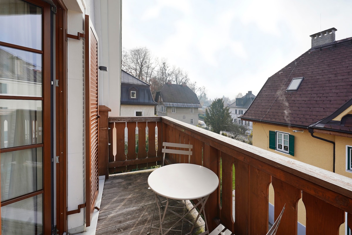 A private balcony is displayed, featuring a round table surrounded by two chairs. Wooden railings add a rustic touch, while neighboring rooftops and trees can be seen in the background. The area is bright, suggesting a pleasant outdoor space for relaxation.