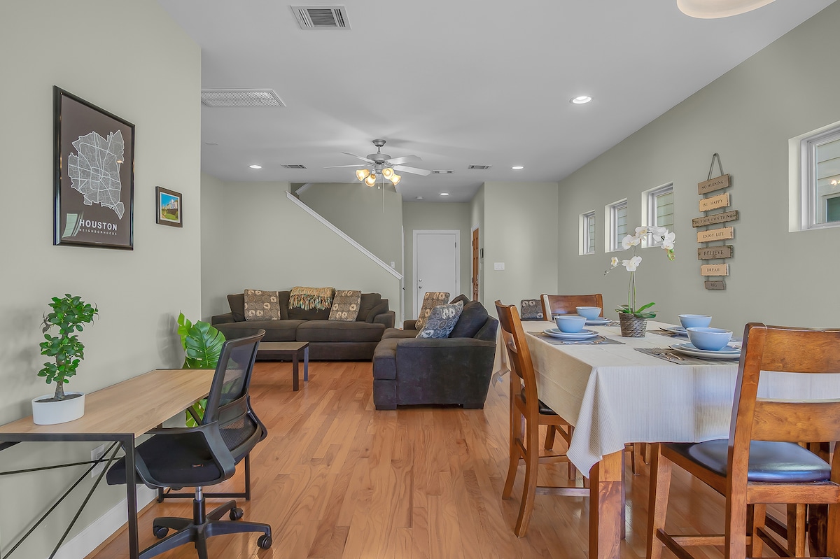 An inviting open-concept living and dining area showcases natural wooden floors. A spacious couch with decorative pillows sits opposite a dining table set for four. A small desk and potted plant are positioned in the corner, while light streams through the multiple windows.