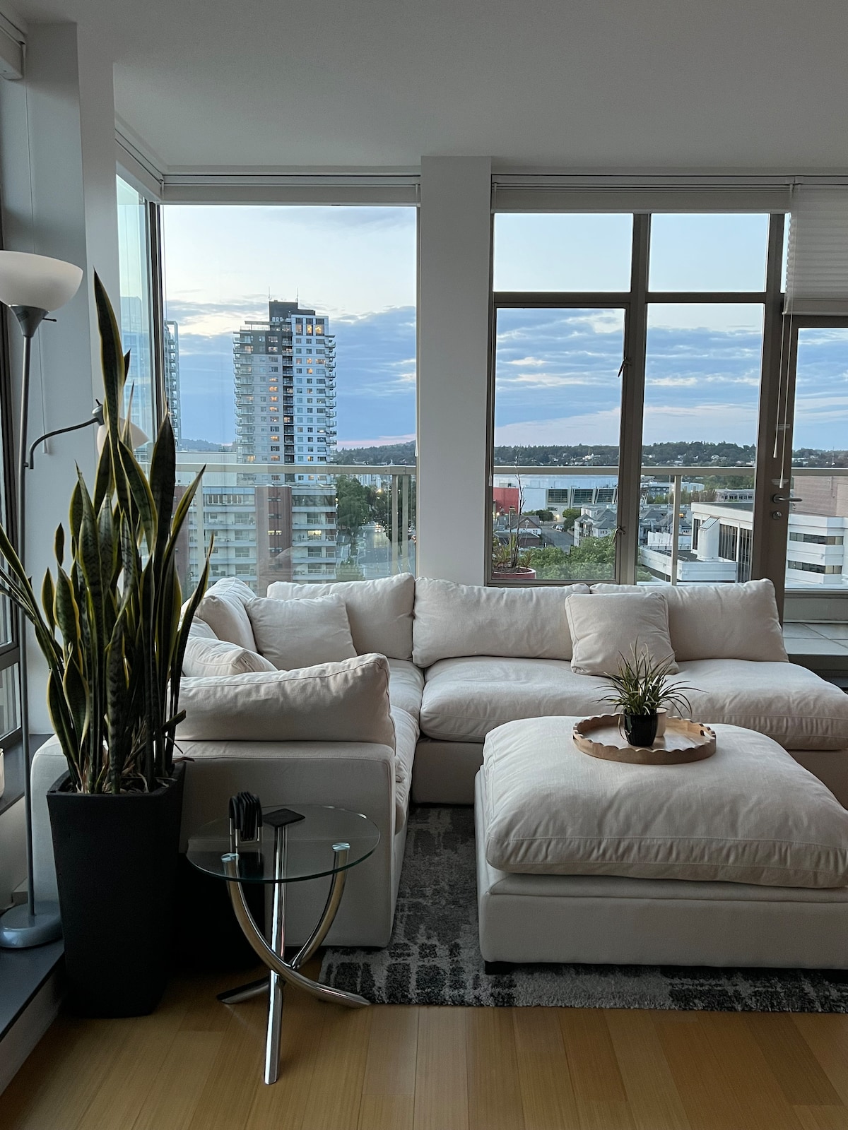 A modern living area is showcased with a plush sectional sofa in light fabric, accompanied by a glass side table. A potted plant adds a touch of greenery beside the window, where city views and a partially cloudy sky are visible.
