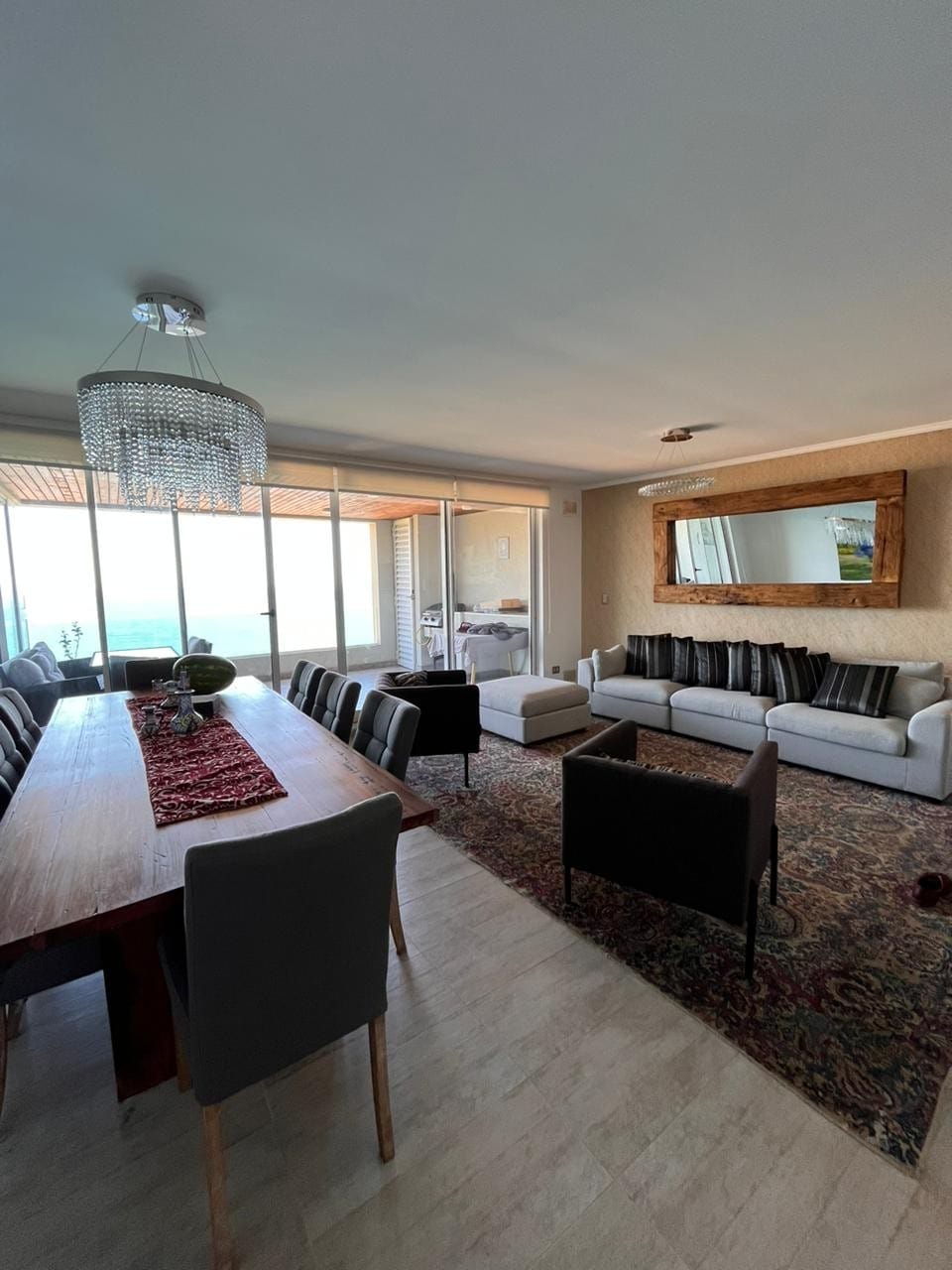 A spacious living and dining area is visible, featuring a large dining table surrounded by grey and black chairs. A comfortable seating arrangement is present, facing a wall-mounted mirror. Natural light floods the space through expansive glass doors, leading to a scenic view.