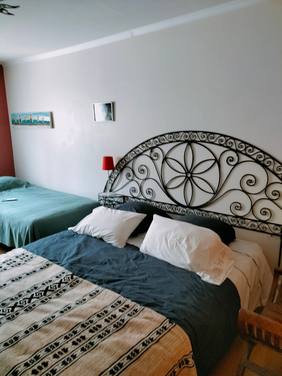 A spacious sleeping area features two beds, one with an ornate headboard and a decorative blanket. A bedside lamp provides soft lighting. Wall art and a small artwork are displayed above the beds, adding subtle decoration to the inviting space.