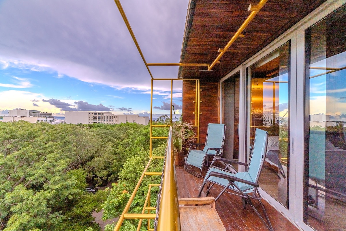 A private balcony is accessed from the living area, featuring two lounge chairs. Lush greenery is seen in the foreground, with a view of city buildings in the background. The sky exhibits varying shades of blue and purple, indicating early evening.