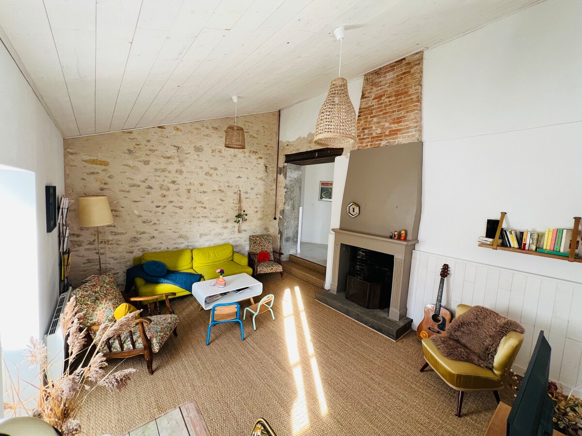 A cozy living area features a vibrant yellow sofa contrasting with floral patterned chairs. A small table is positioned in the center, with a window allowing natural light and a chimney adding warmth. Light-colored walls and rustic exposed brick enhance the inviting atmosphere.