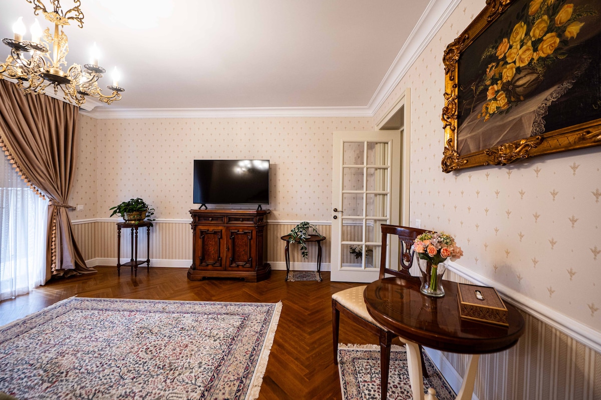 A finely decorated living area features elegant wallpaper and a wooden side table with flowers. A large television is mounted on the wall opposite a stylish cabinet. Natural light enters through a window, with light drapes gently framing the view.