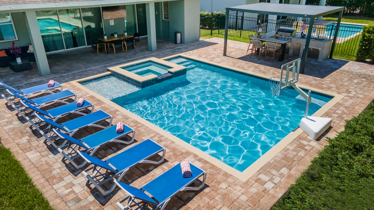 The outdoor area features a spacious swimming pool with an attached spa. Lounge chairs are arranged around the pool, complemented by a shaded dining space equipped with a table and propane grill. A basketball hoop is positioned at the pool's edge, enhancing recreation options.