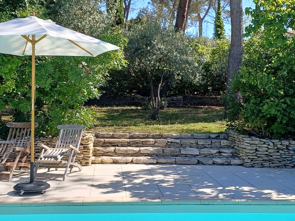 A refreshing swimming pool is bordered by natural stone walls, leading to a lush garden area. Two wooden loungers are positioned under a large umbrella, providing a relaxing spot amidst the greenery. Dappled sunlight filters through nearby trees.