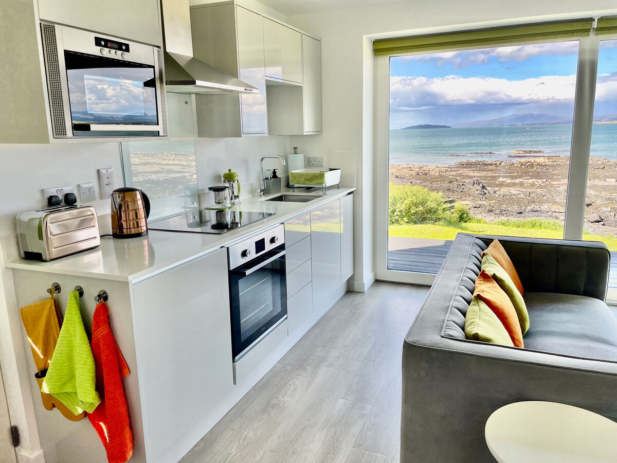 A modern kitchen is equipped with sleek appliances, including an oven, microwave, and a counter fridge. Colorful kitchen towels hang beside a toaster. A comfortable seating area features a gray sofa adorned with bright cushions, and large windows offer views of the serene waterfront.