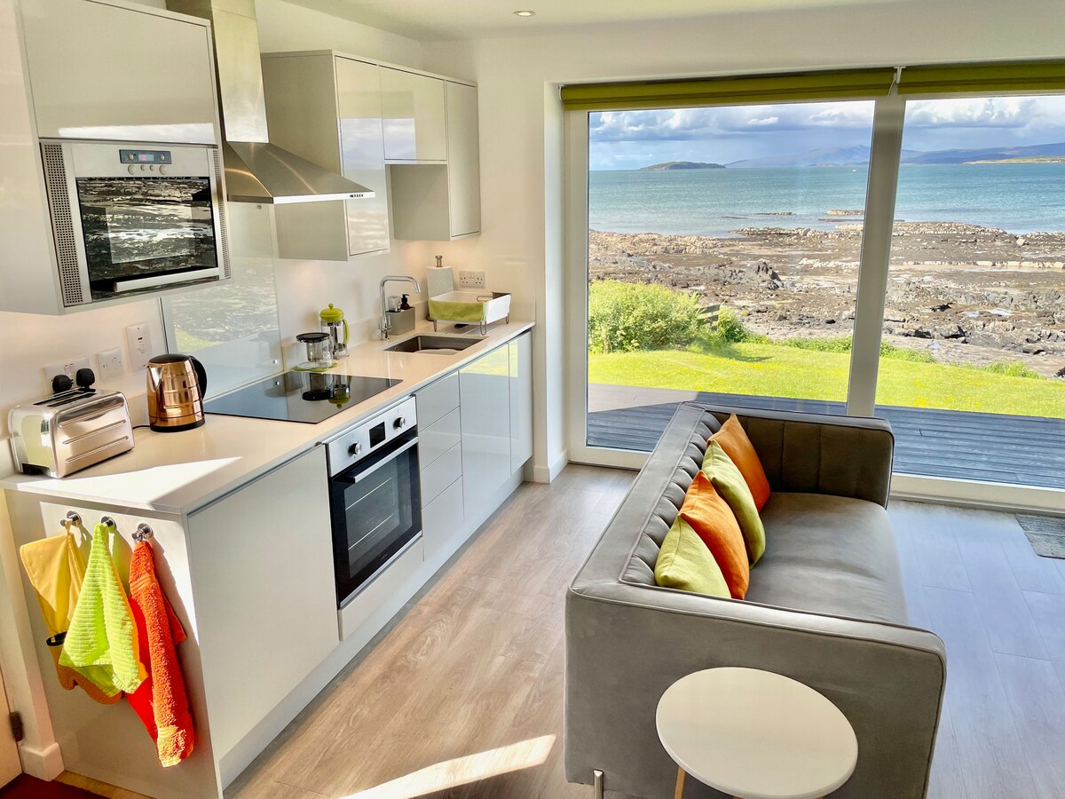 A bright and modern kitchen area features sleek cabinetry and stainless steel appliances. Natural light floods the space through large windows overlooking the shoreline. A comfortable sofa with colorful cushions complements a small round table, creating an inviting setting for relaxation.